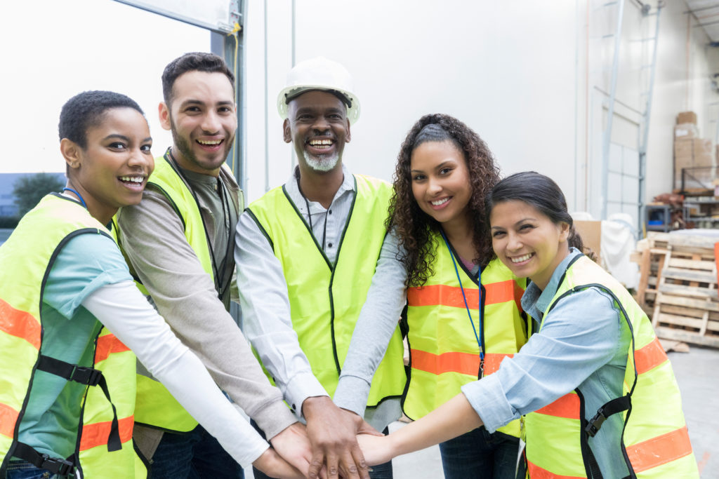 Confident diverse group of distribution warehouse employees have their hands together in unity. They are smiling at the camera.